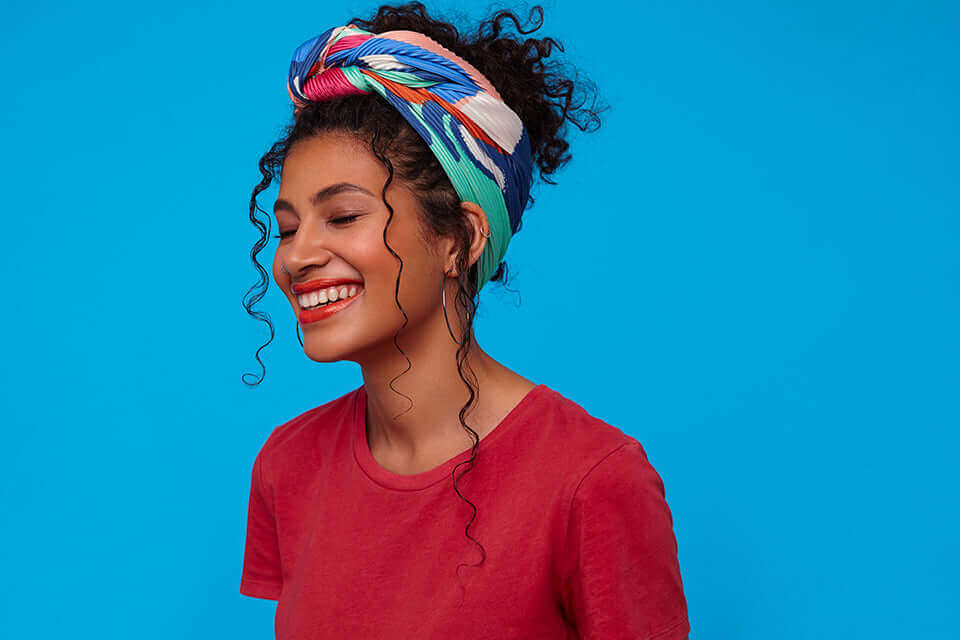 Femme souriante avec un turban coloré et des cheveux bouclés sur fond bleu.